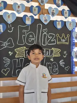 Niño del colegio posa frente al tablero decorado con corazones y fotos de compañeros