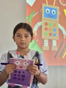 Niña del colegio sostiene un robot de cartulina rosa y morada que elaboró en clase