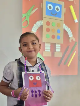 Estudiante sonríe mientras muestra su robot de papel frente a un mural de robótica